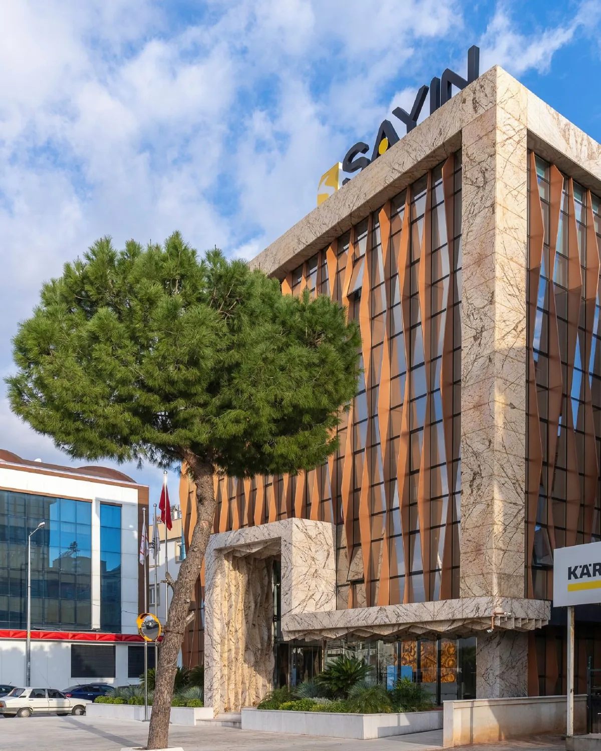 SAYIN binası — mermer çerçeveli, bakır lamelli cam cephe ve girişteki çam ağacı